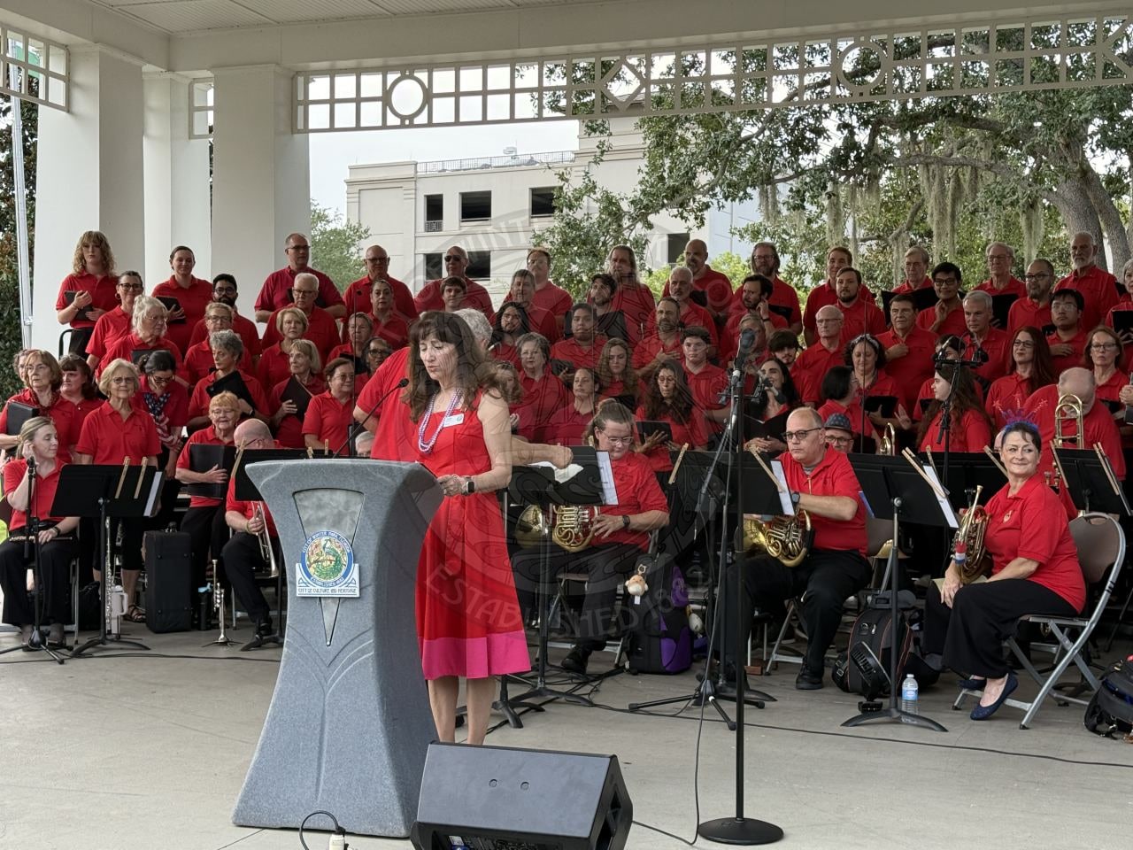 A woman in a red dress speaks at a podium with a large choir and band in red shirts behind her on stage.