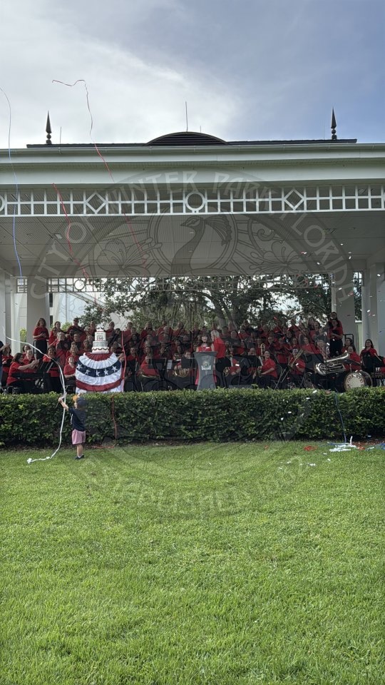 A child with streamers stands on grass before a band playing under a white gazebo on a cloudy day.