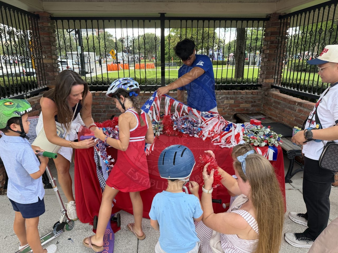Adults and children decorate with red, white, and blue crafts at an outdoor covered table near a metal fence.