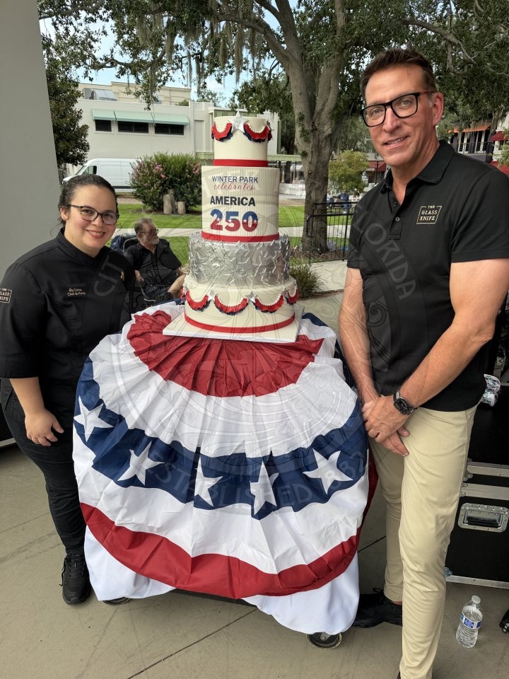 Two people stand by a patriotic cake and star-spangled cloth, celebrating America’s 250th anniversary outdoors.
