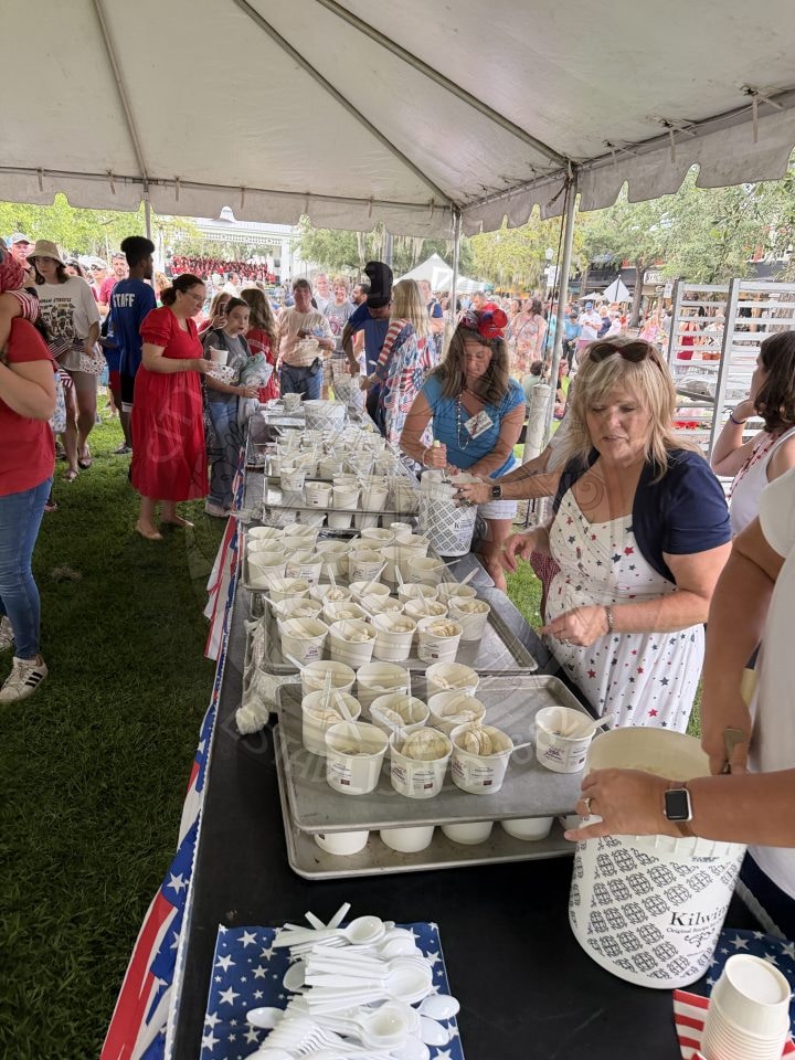 People serving and receiving cups of ice cream at a crowded outdoor event under a tent.
