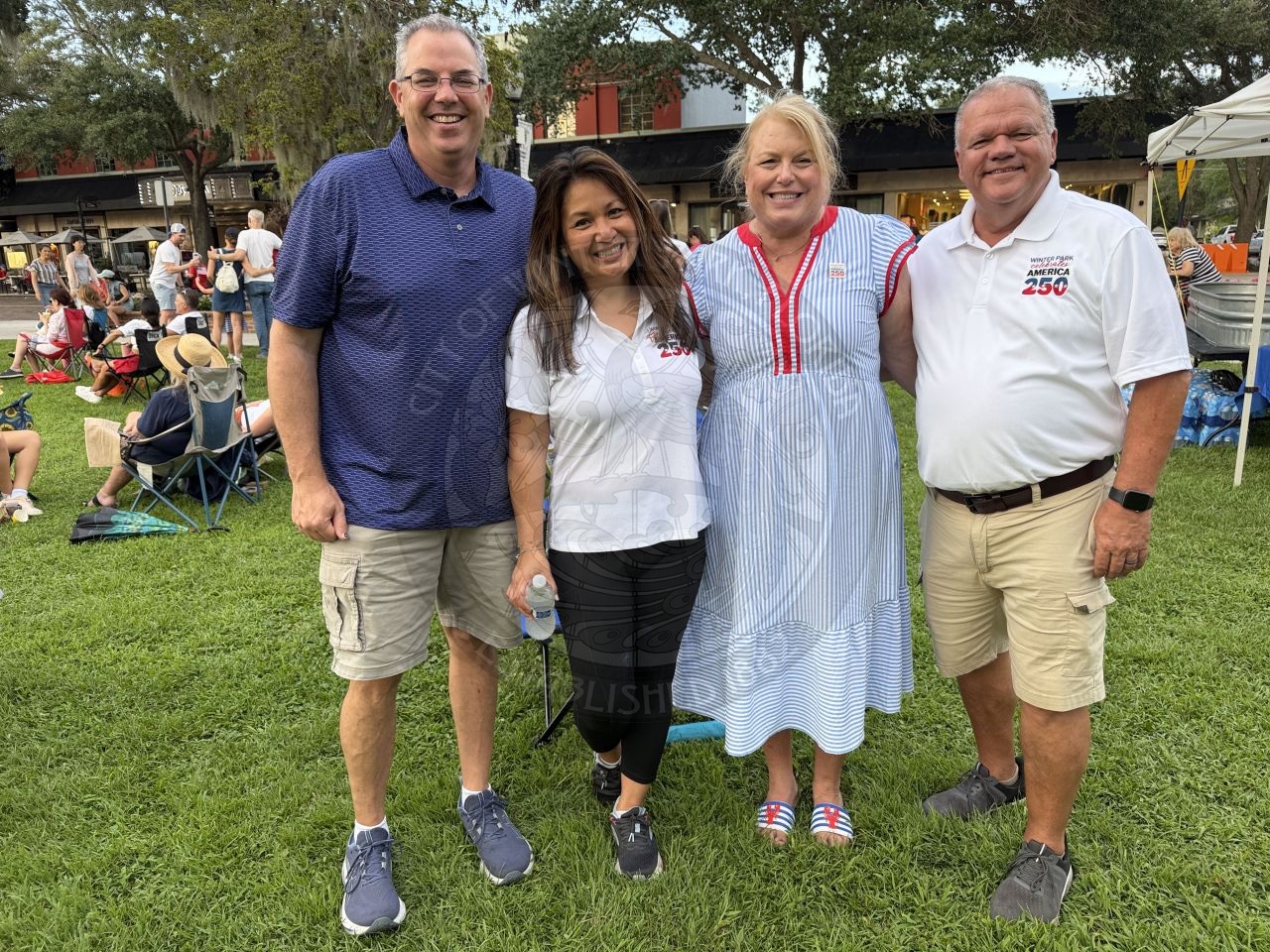 Four adults standing on grass, smiling at an outdoor event with people and tents in the background.