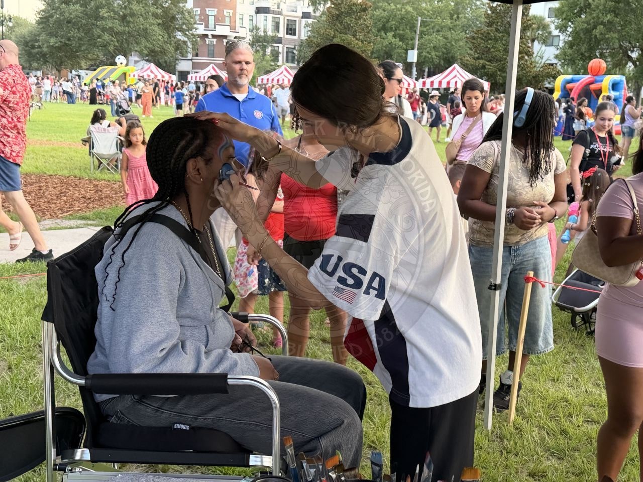 A woman paints a childs face at an outdoor festival, with people and tents visible in the background.