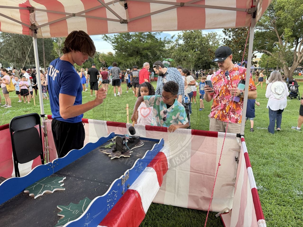 Two boys play a carnival game under a red and white tent; a man in blue shirt watches and people are in the background.