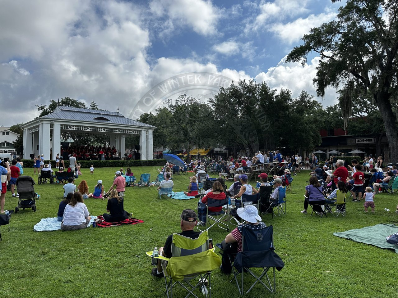 people sitted in lawn chairs and and on blankets in the grass area listening to an orchestra perform.