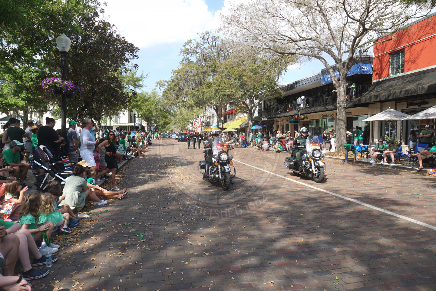 2 police motorcylces riding next to each other waving to the people in the sidewalks