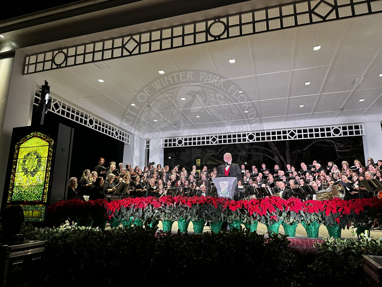 the director of Bach standing on the stage behind a podium with the choir behind him waiting to perform. To the left of the stage is a yellow/green stained glass window with a wreath on it.