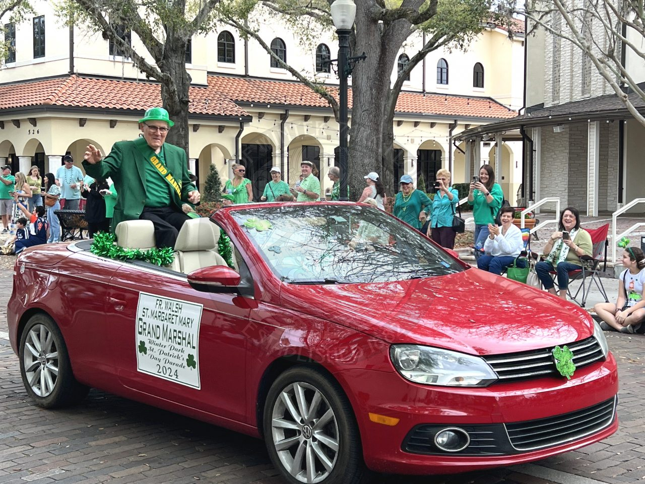 A red car with a man sitting in the back seat/trunk of car waving to people as the car drives down Park Avenue