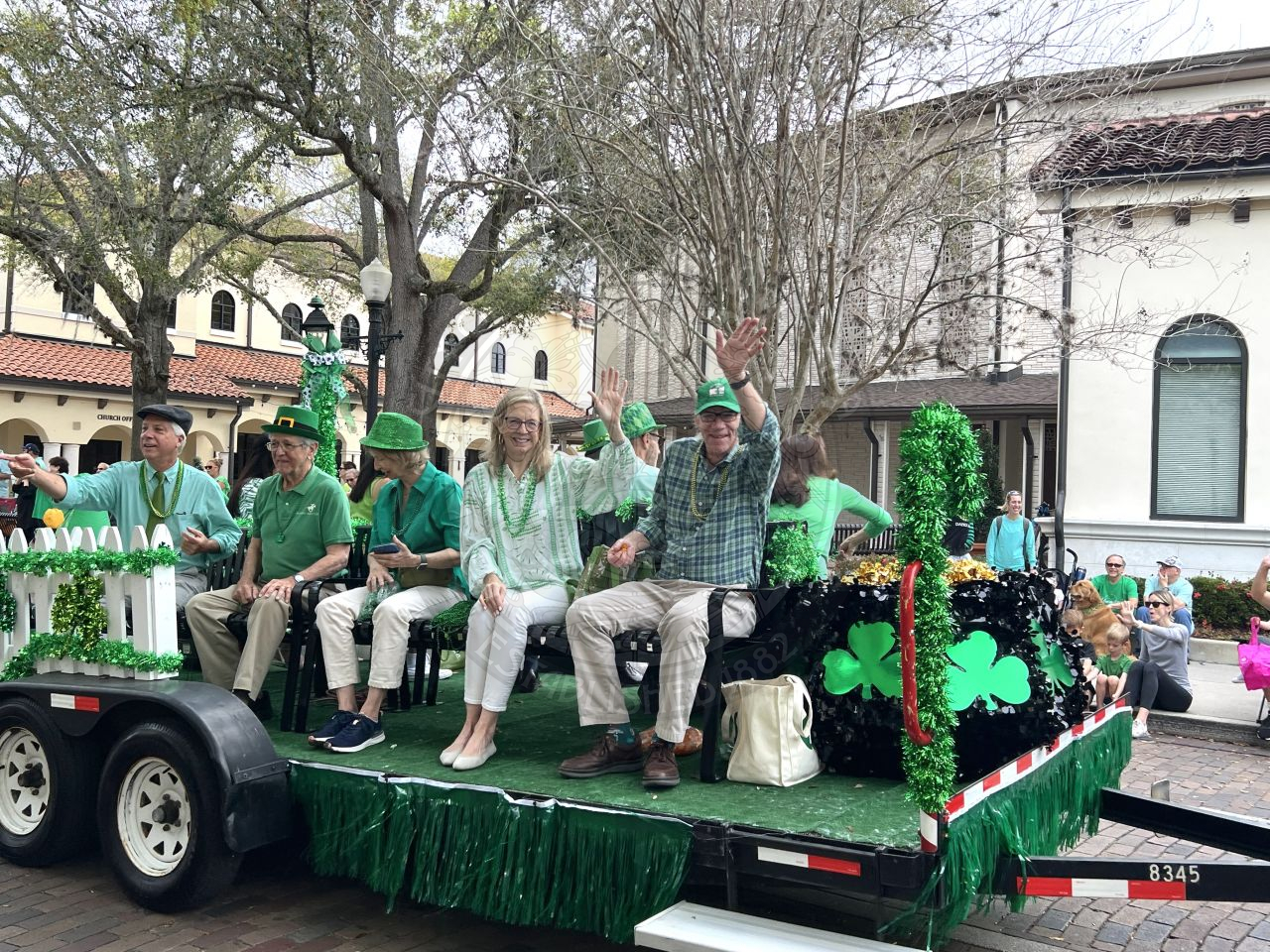 Mayor and commissioners sitting on a bench on a a loat with green st. patrick's day decorations thrwing out beads to the people watching the parade.