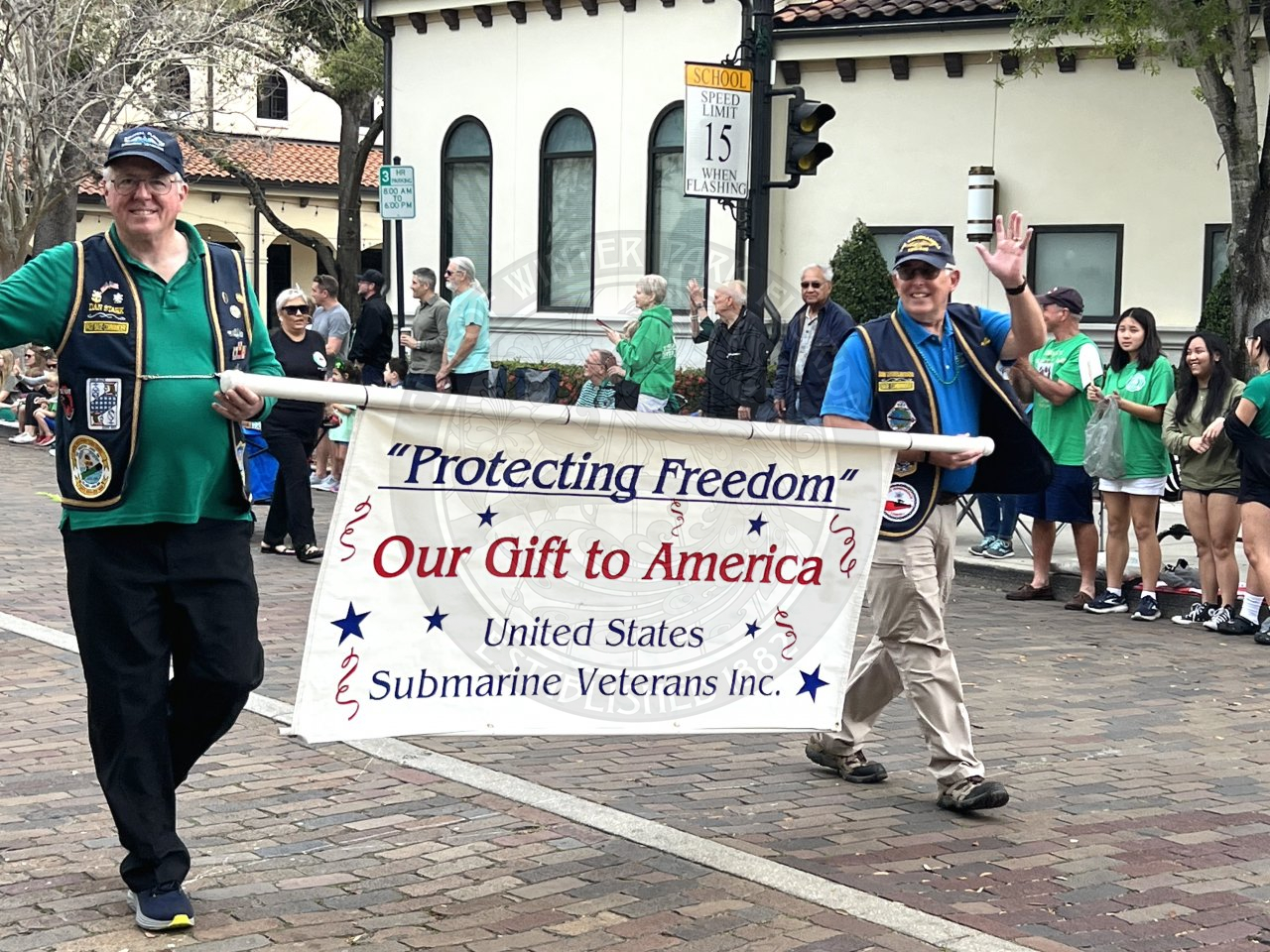 two men holding a banner that says "Protecting Freedom Our gift to America United States Submarine Veterans Inc." The men are holding each end of the pole that has the banner on it.