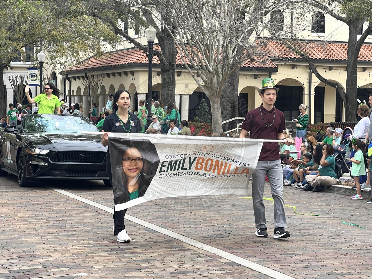 2 young kids holding a sign for politican Emily Bonilla. Behind them is a mustang where Emily is sitting in the car waiving to everyone on the sidewalks.