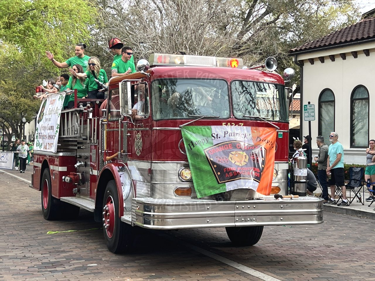 The front view of a firetruck with people at the top of it wearing green shirts and waving to the people watching the parade on the sidewalks.