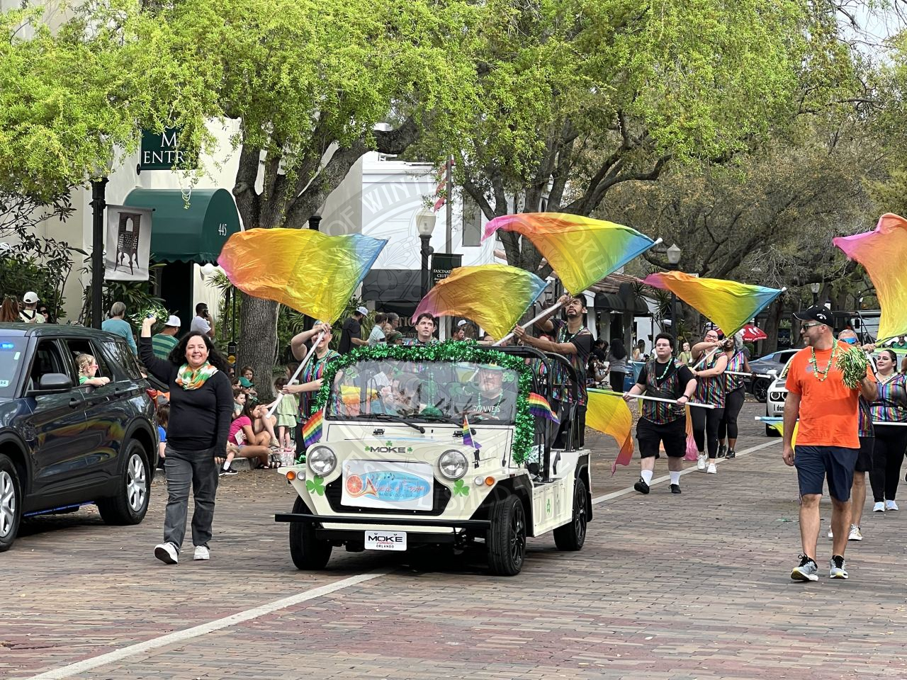 A moke driving down Park Avenue with people marching down behind the car with people swinging flags that are rainbow colored.