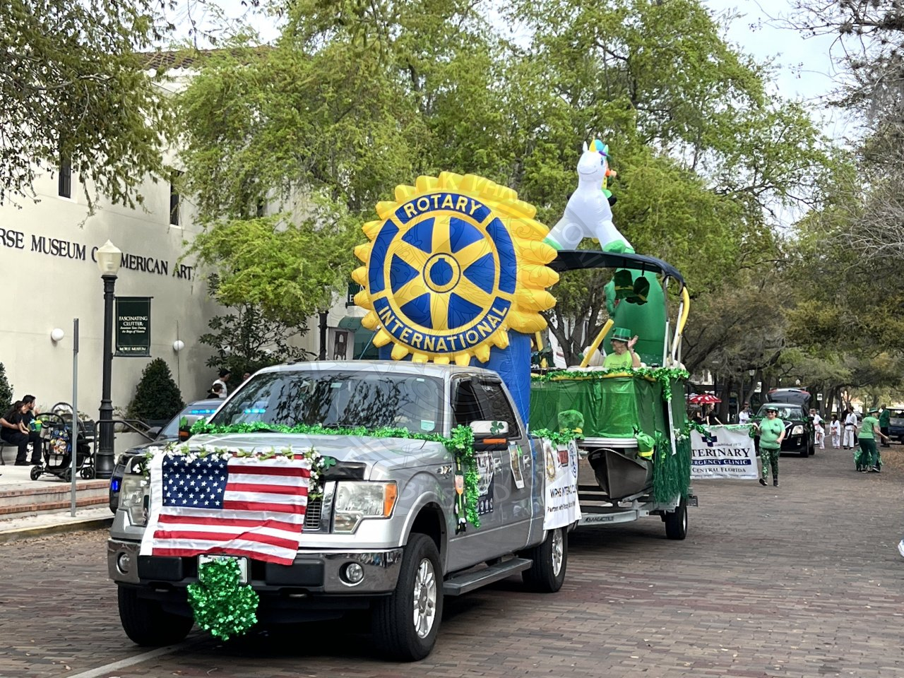 silver truck driving down Park Avenue with St. Patrick's day decor with a green boat behind the truck.