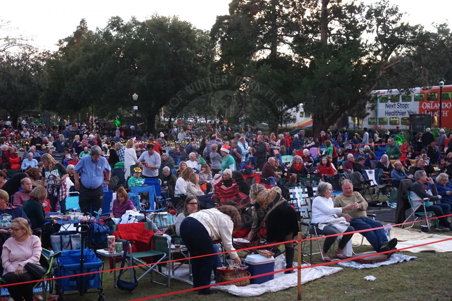 People sitting in chairs and on picnic blankets in the park enjoying food and drinks while waiting for the concert to start.