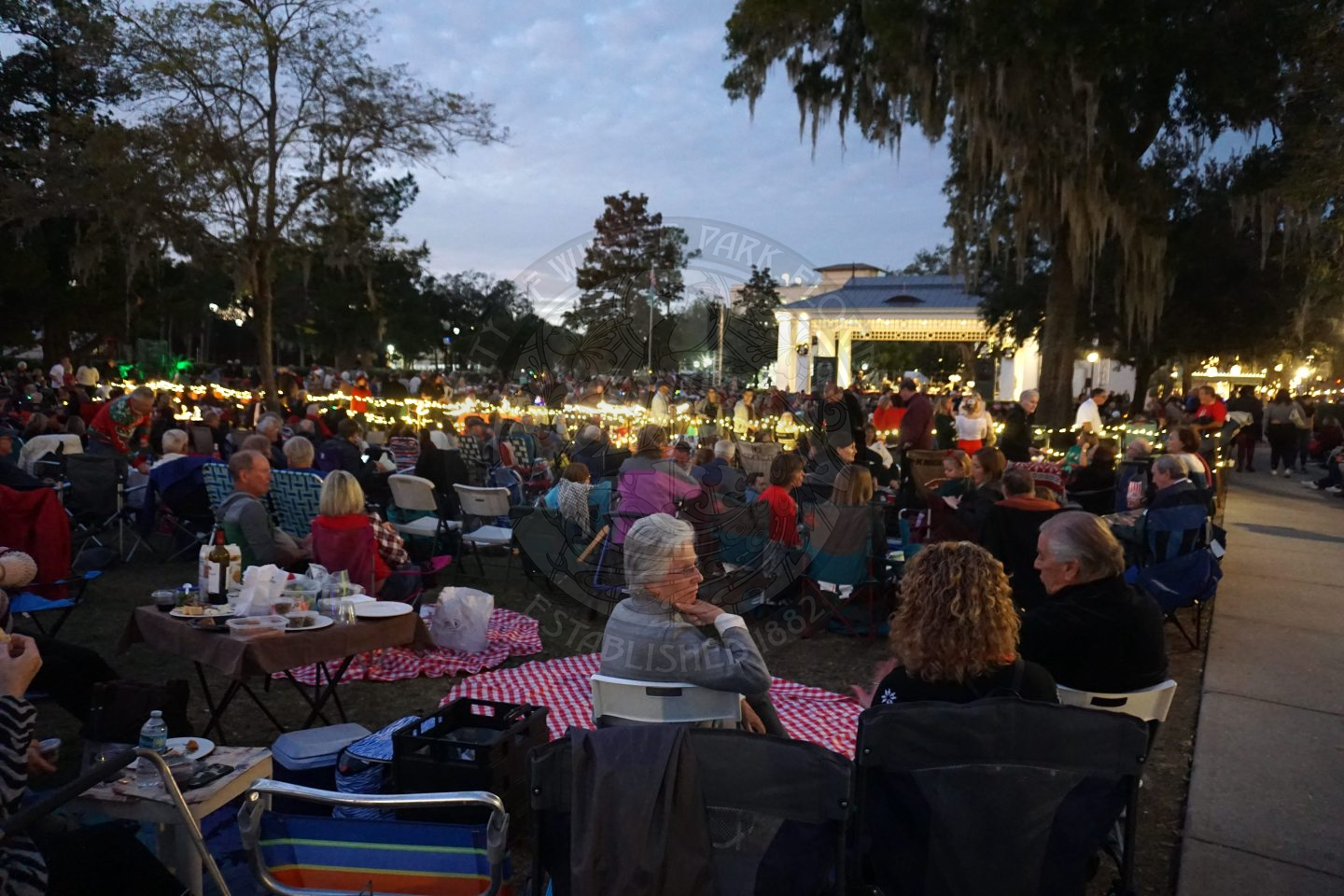 a crowded group of people sitting down in chairs or blankets facing the stage where there is a symphonic band playing.
