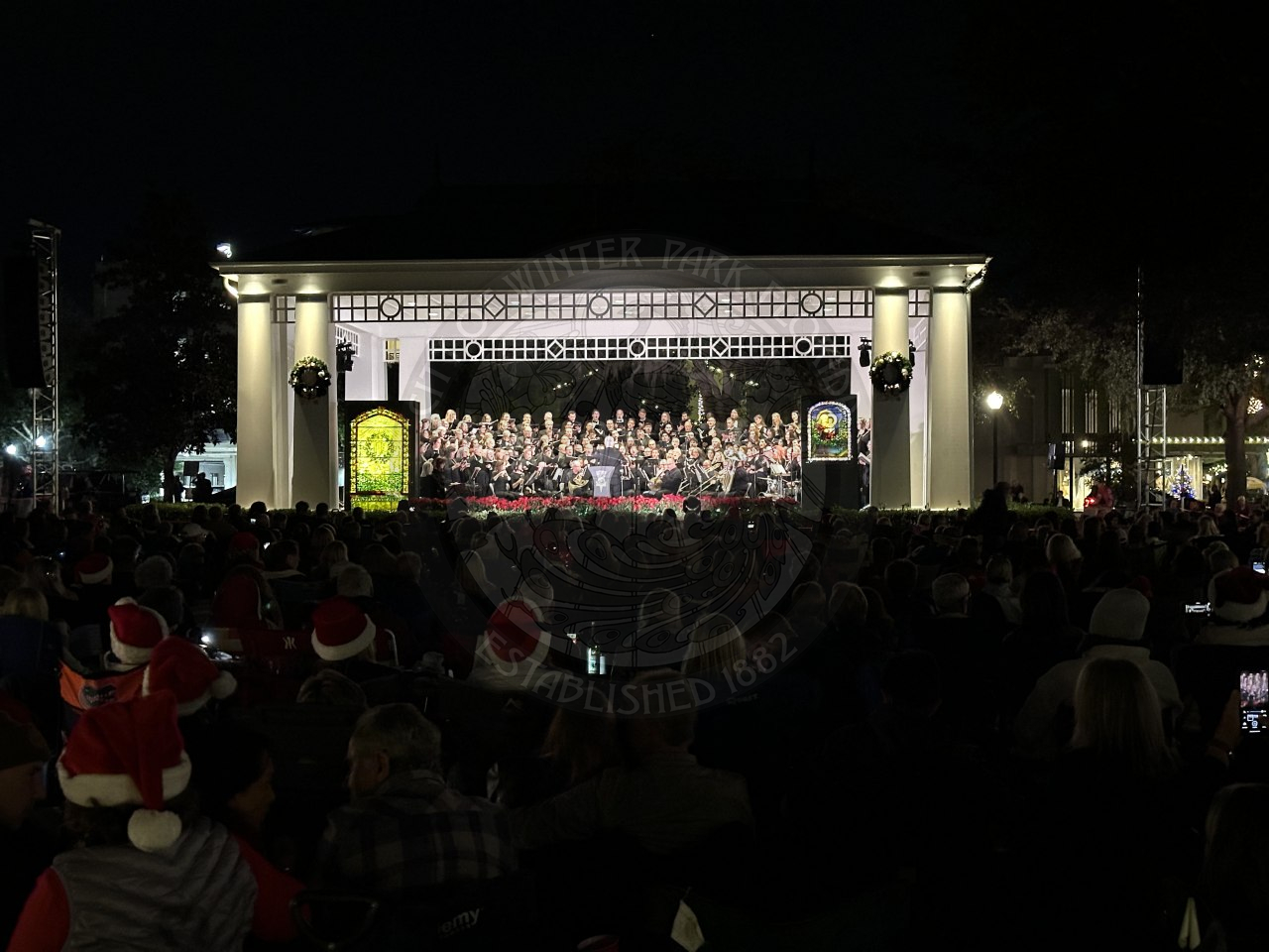 An outside stage with two tiffany stained glass windows on the left and right side of the stage. In the middle is a symphonic band sitting and playing their instrumetns to perform a concert.