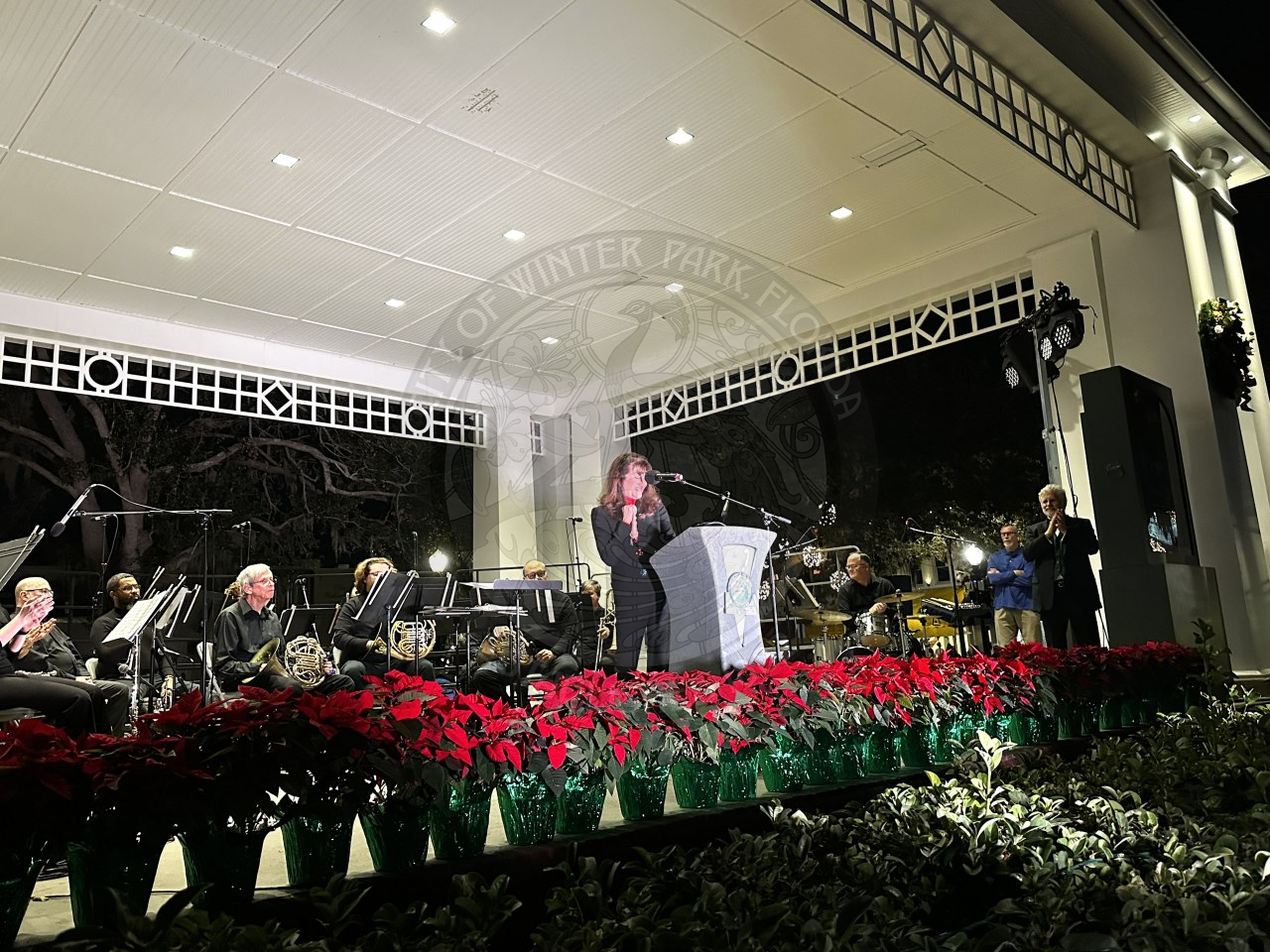 A woman standing behind a podium talking into a microphone into the crowd. There is a row of poinsettas lining the stage with a band in the back of the stage behind the lady speaking.