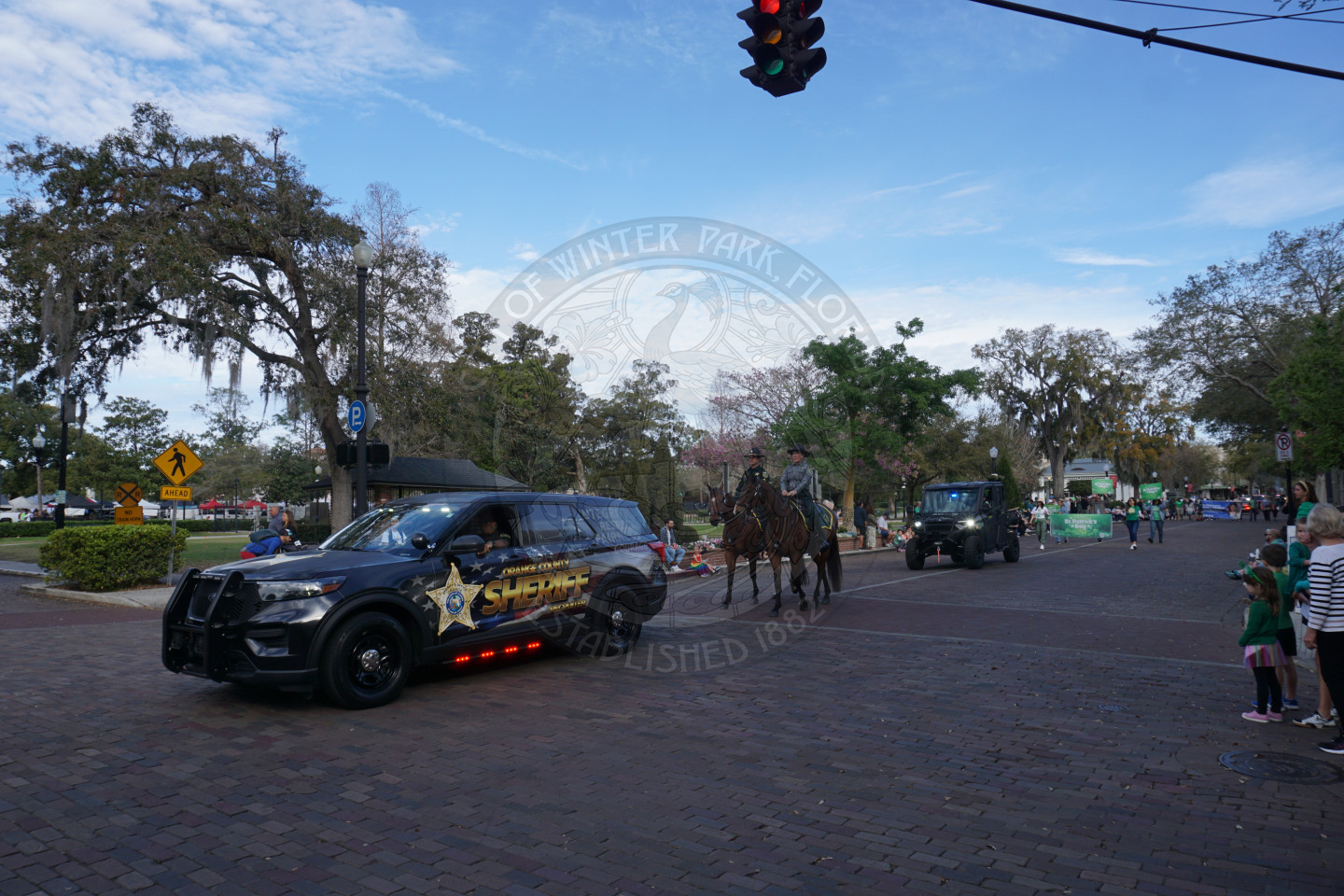 Orange County Sheriff's car with two horses behind the car traveling on a brick street.