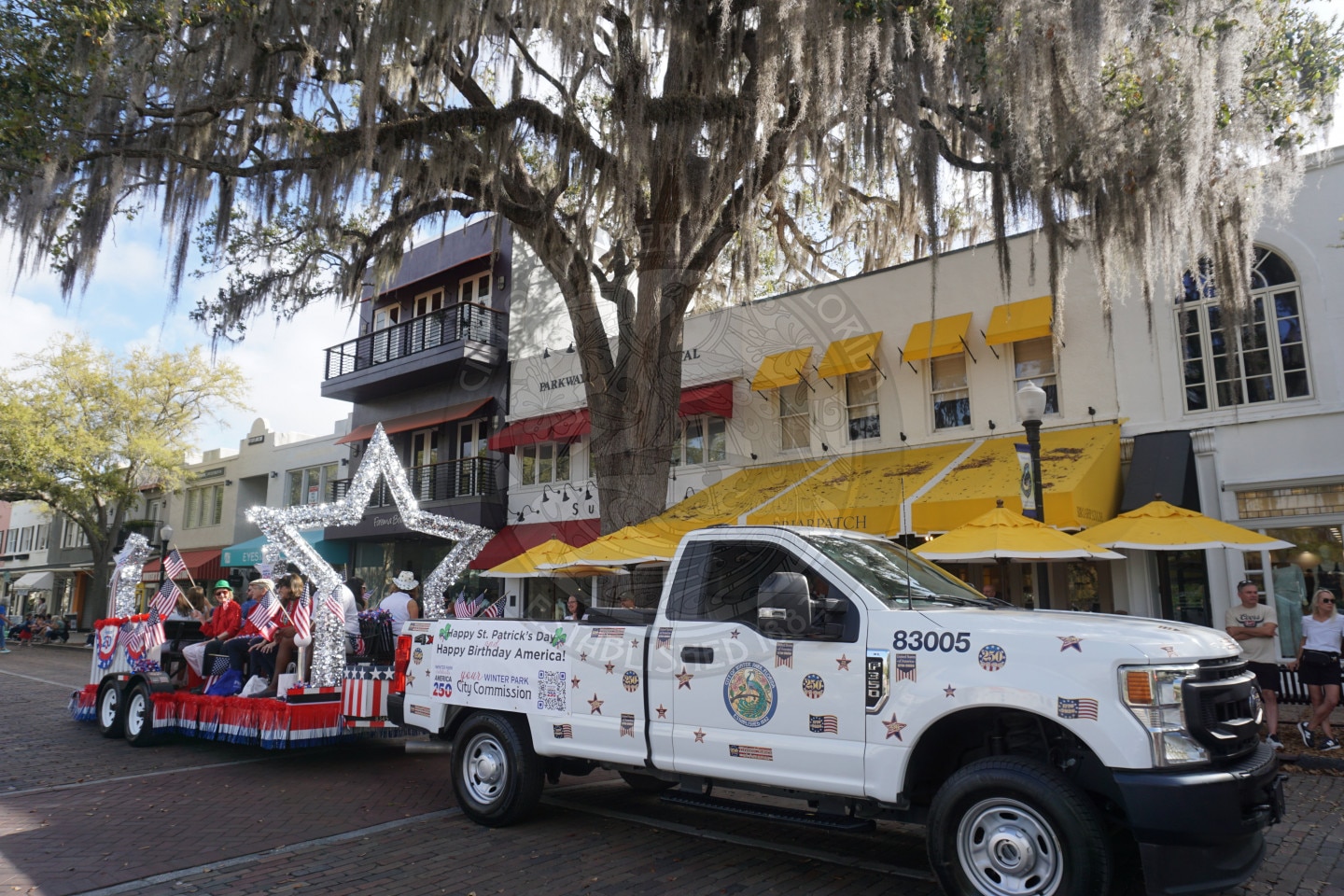 A decorated white truck towing a patriotic float with people in festive attire passes shops and cafes under large, moss-covered trees during a parade on a sunny day.