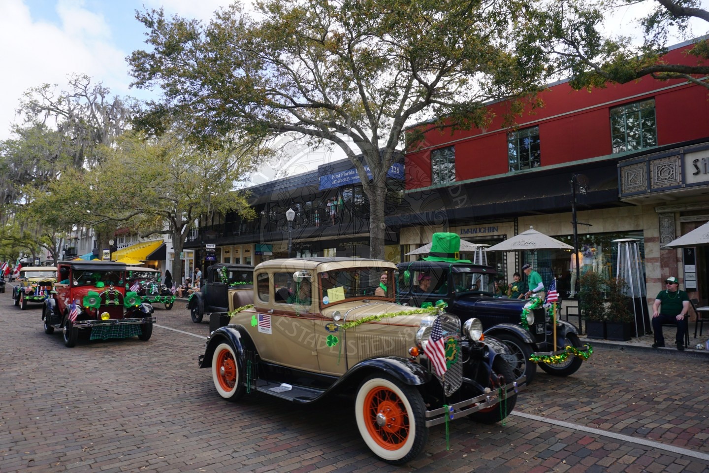Vintage cars decorated with shamrocks and American flags drive down a brick street lined with trees and shops during a festive parade, possibly for St. Patrick’s Day. People sit and stand along the sidewalk watching.