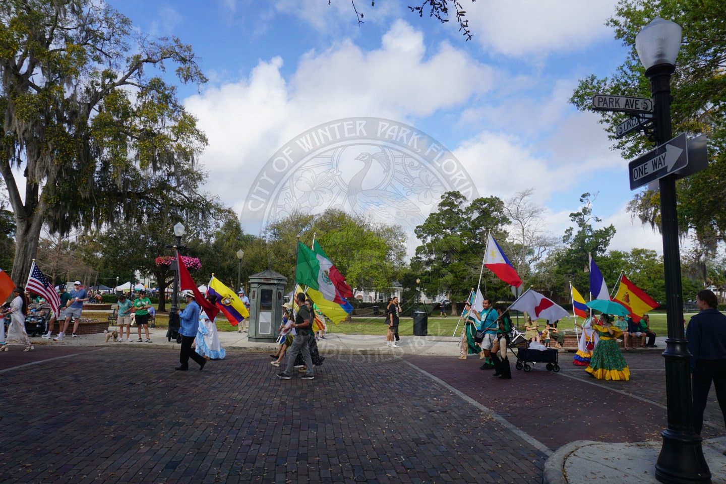 People carrying colorful international flags walk through a brick-paved street at a park festival in Winter Park, Florida. Trees and street signs frame the lively outdoor scene under a partly cloudy sky.