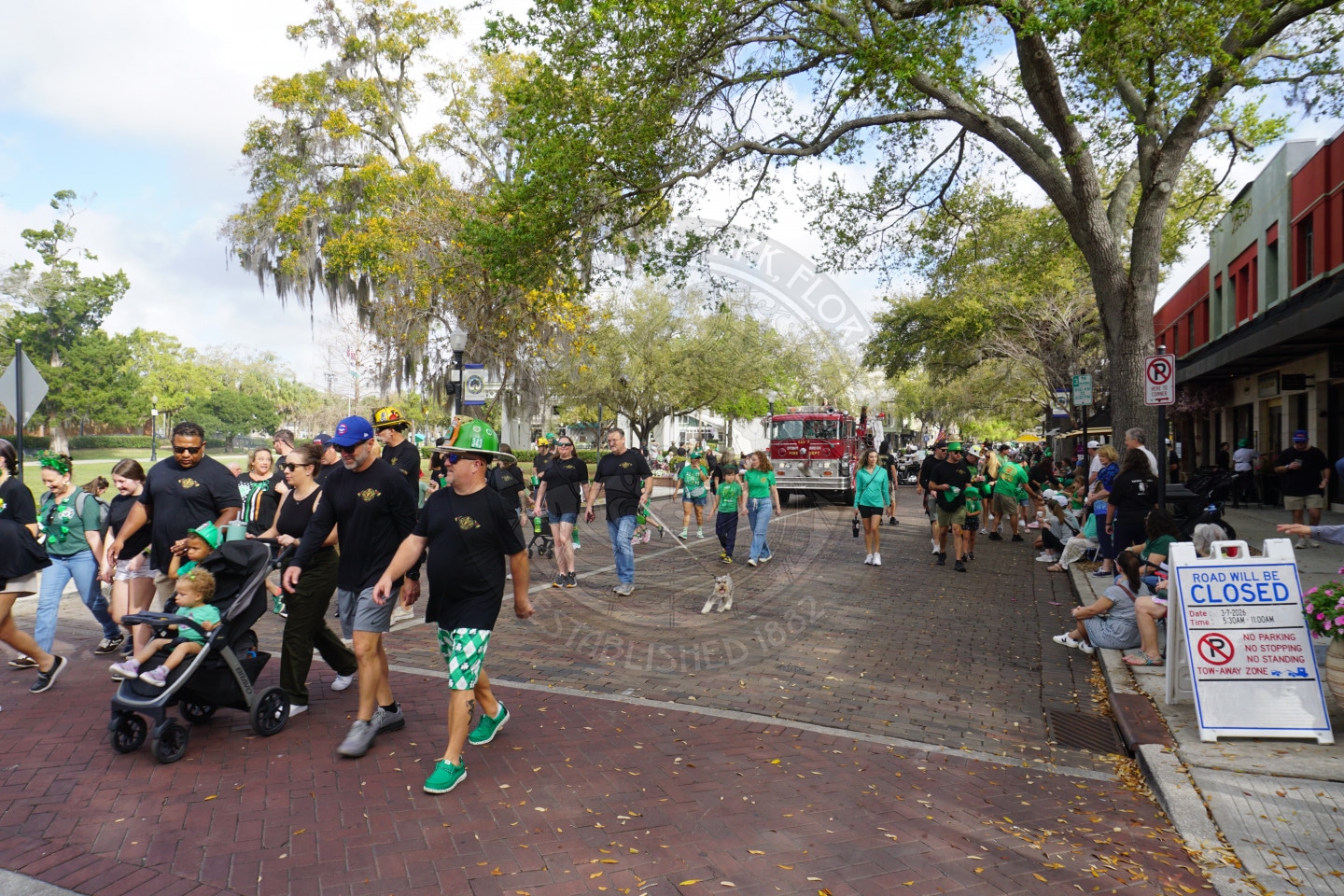 A group of people, many wearing green, walk in a festive parade on a tree-lined street. Some push strollers, others walk dogs. A firetruck is visible in the background.