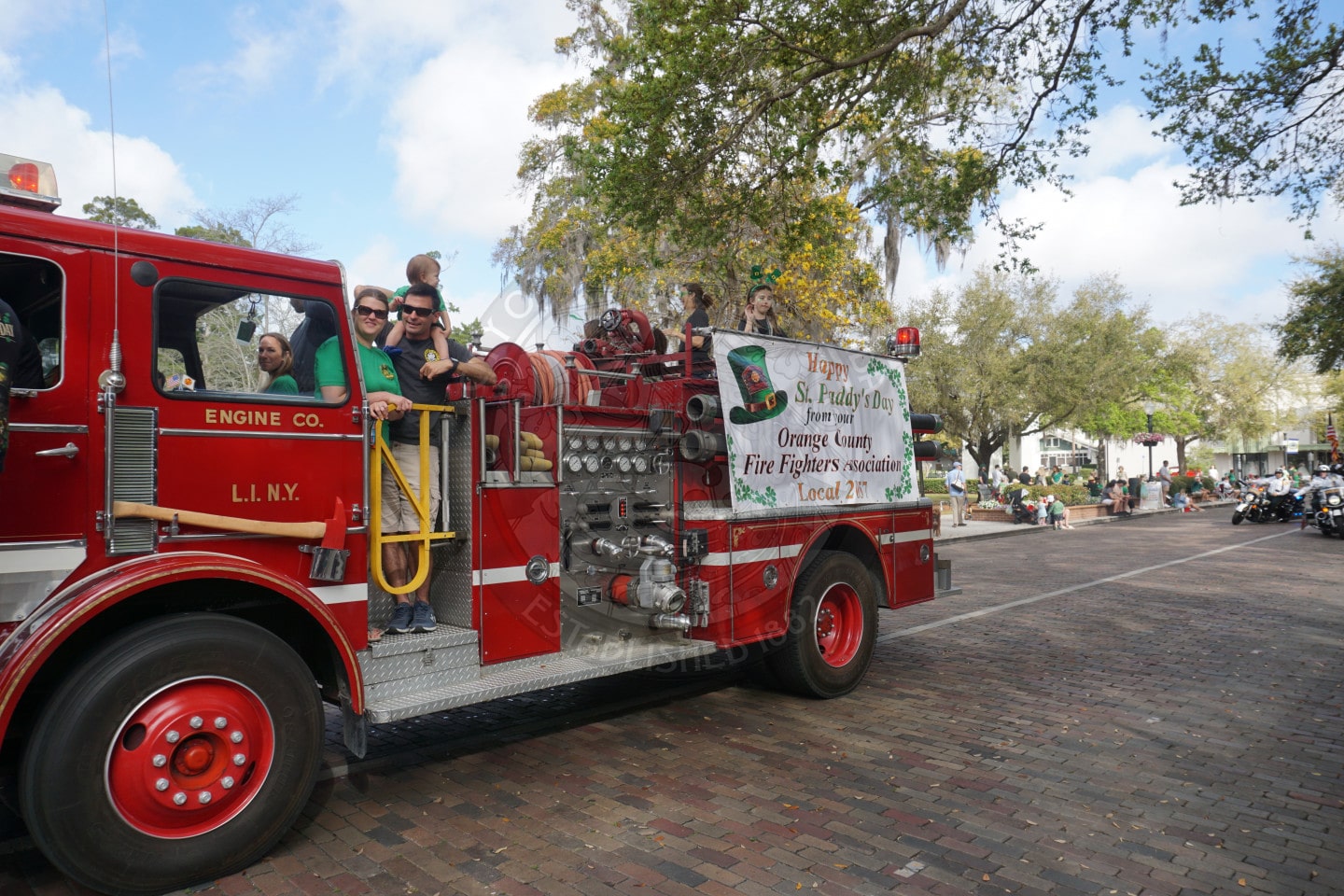 A vintage red fire truck with people in green shirts riding on the side drives in a parade along a tree-lined street, with onlookers sitting and standing on the sidewalk.