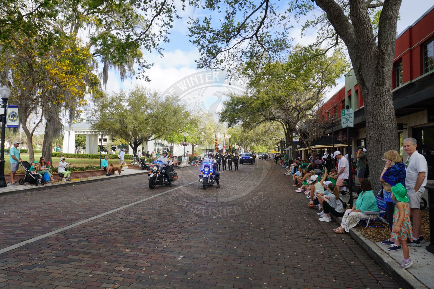 A parade on a brick street with people sitting on both sides, some wearing green. Police on motorcycles lead the parade, followed by people carrying flags. Trees line the street, and buildings are visible on the right.