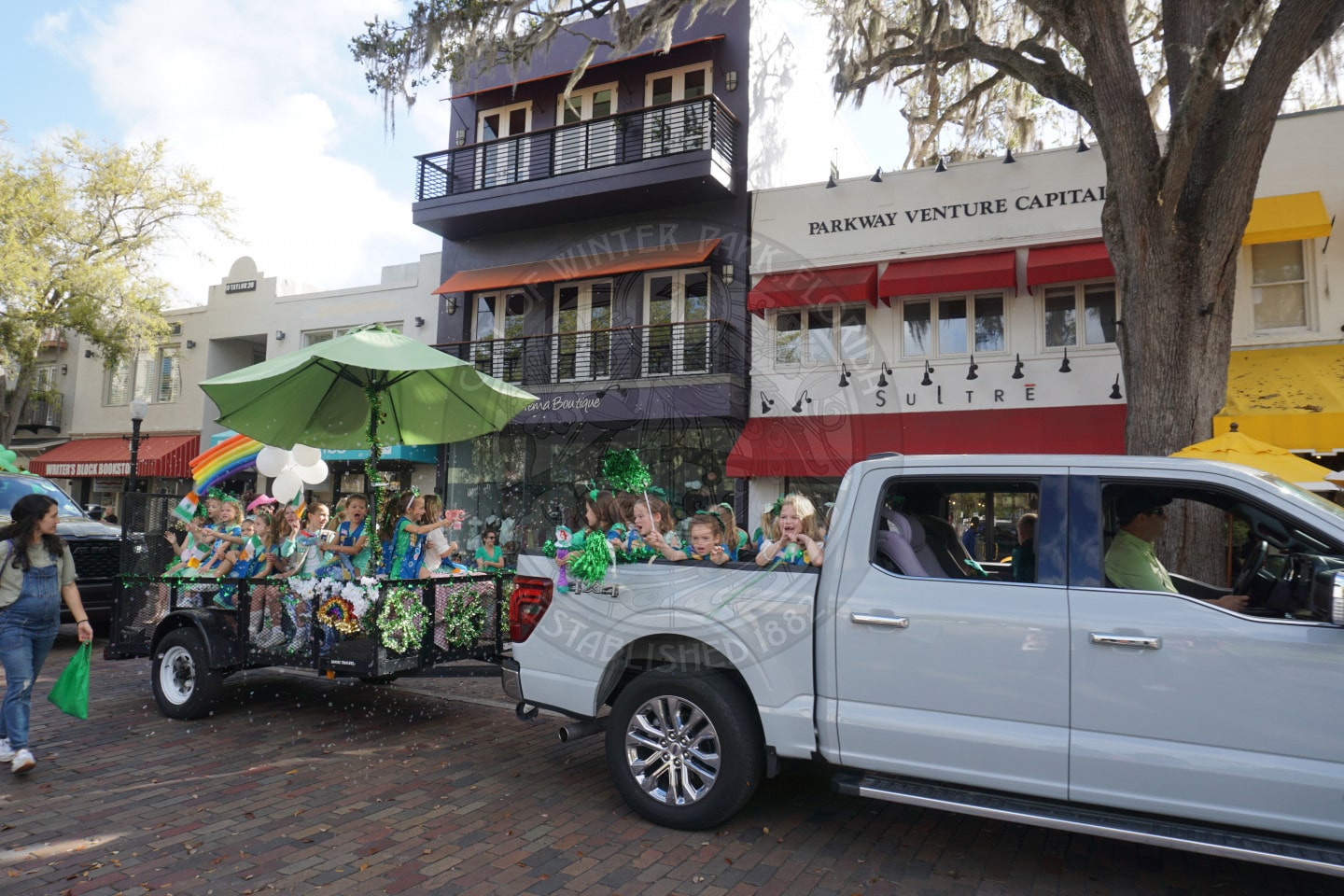 A white pickup truck pulls a decorated trailer with children in green attire, celebrating what appears to be St. Patricks Day, on a brick street lined with colorful buildings and trees.
