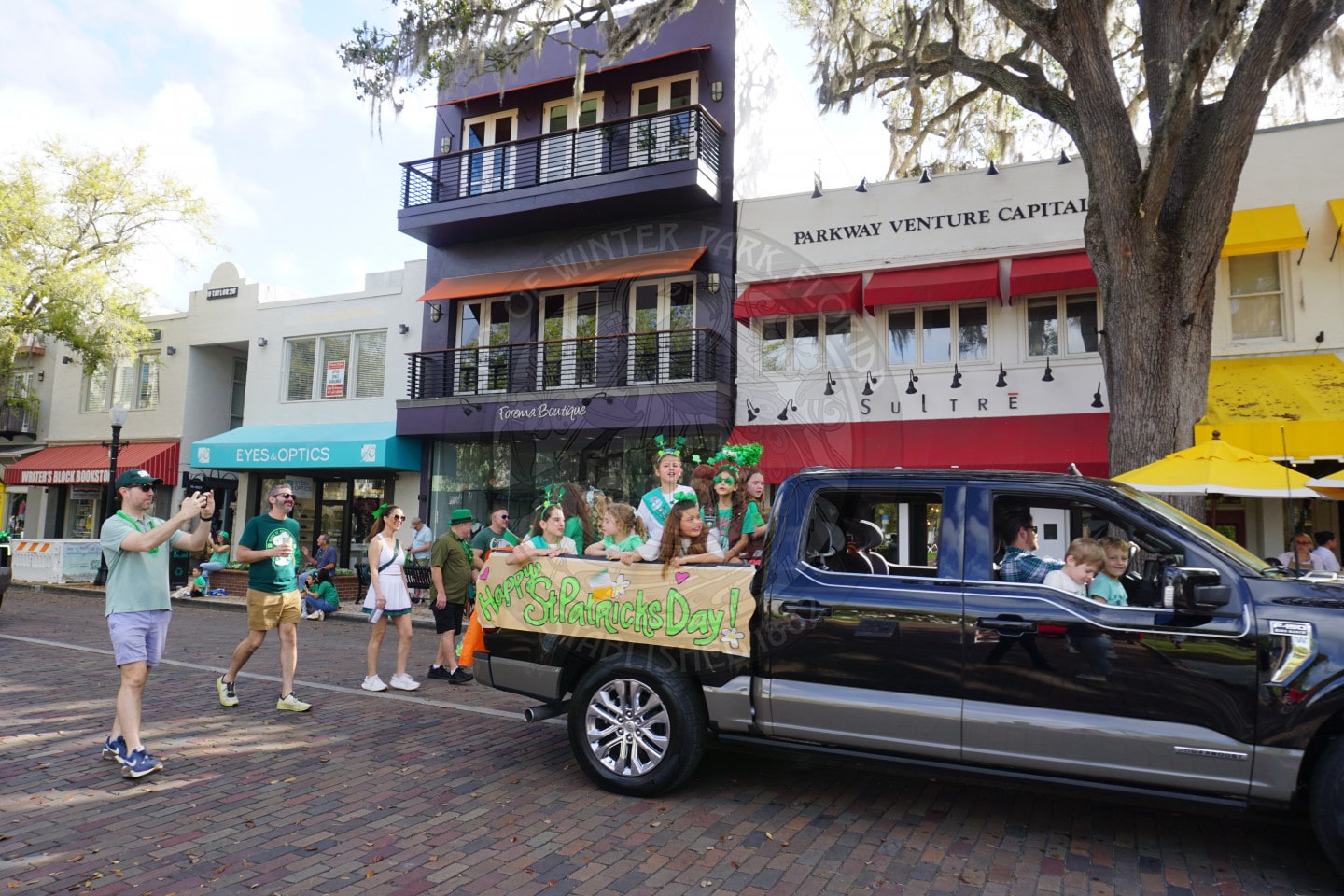 A girl scout troop wearing green celebrate St. Patrick’s Day next to a decorated pickup truck with a banner, on a brick street in front of colorful shops and buildings.