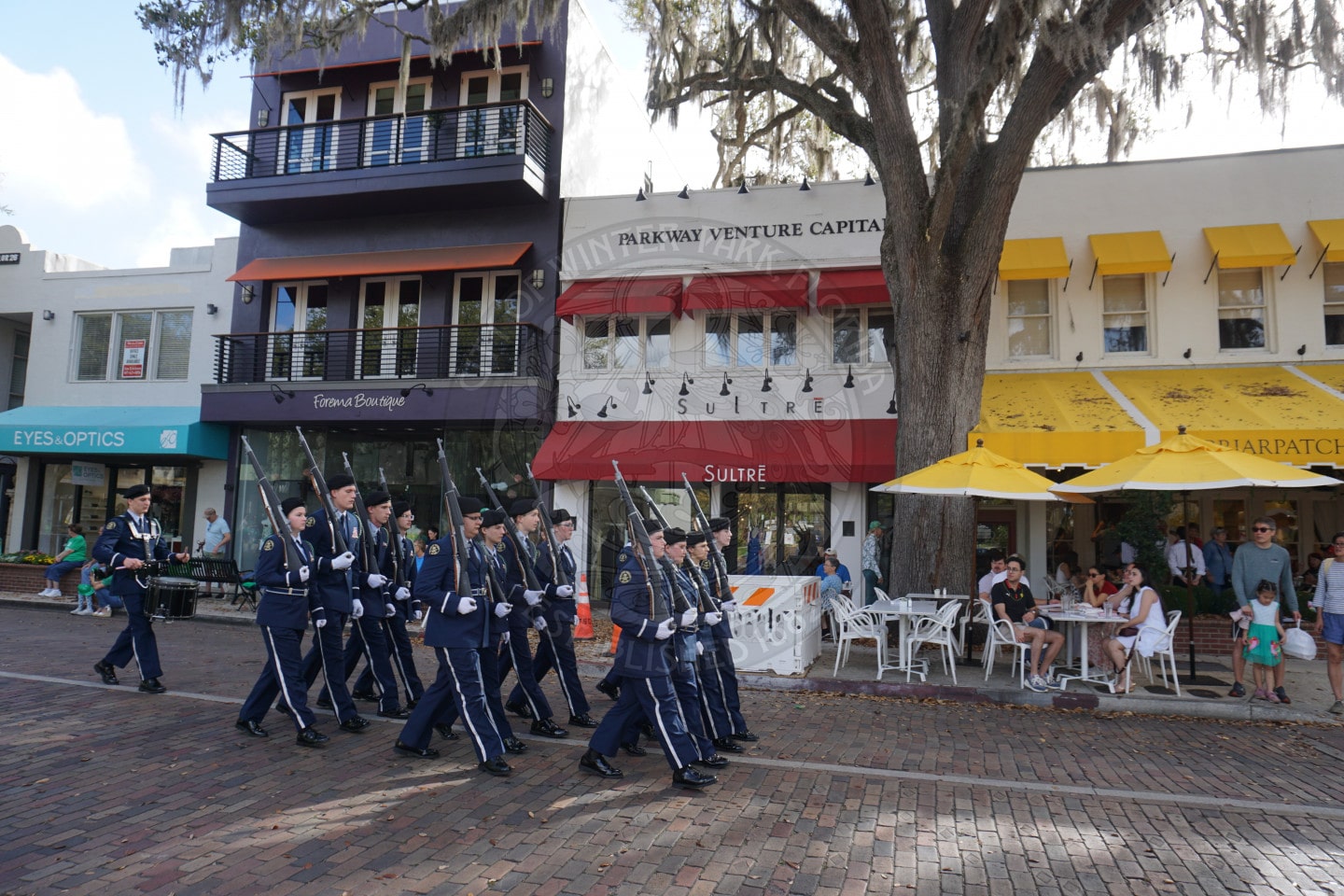 A group of uniformed cadets march in formation with rifles on a brick street past sidewalk cafes and shops, as people sitting outside watch. Trees with Spanish moss hang over the scene.