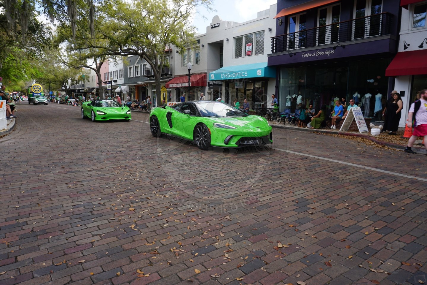 Two bright green sports cars drive down a brick-paved street lined with boutiques, shops, and people walking on the sidewalk under leafy trees.