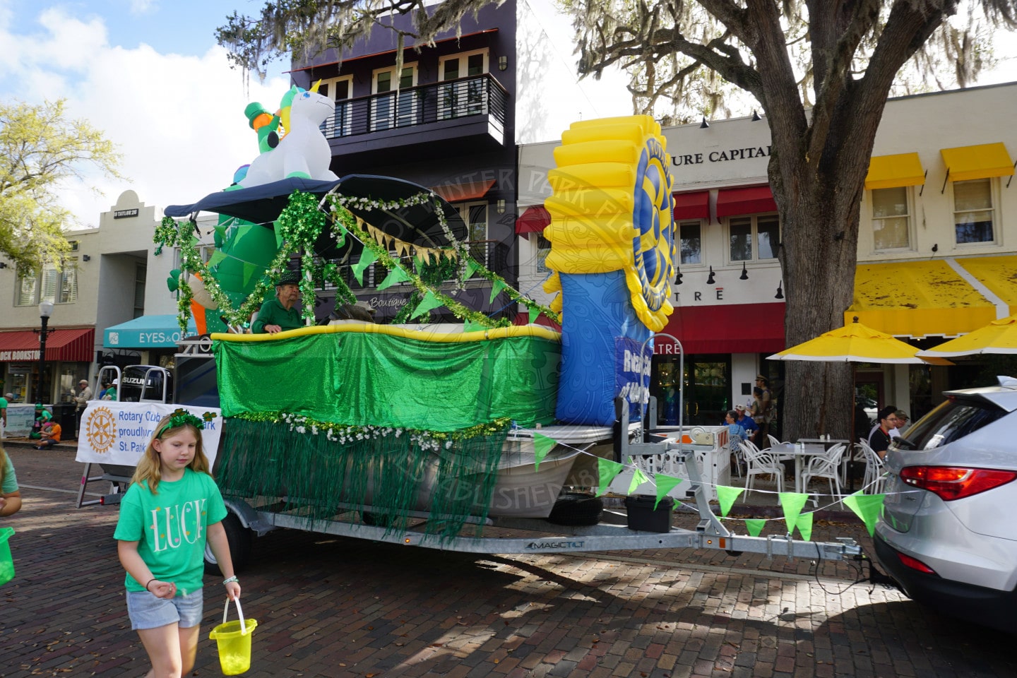 A parade float decorated in green and yellow with shamrocks and Rotary Club symbols moves down a brick street. People in green shirts walk nearby, and outdoor diners sit at tables under large trees and umbrellas.