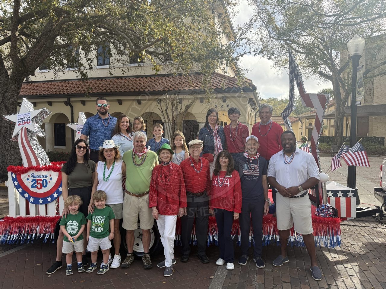 A diverse group of people of all ages smile in front of a patriotic parade float decorated with red, white, and blue bunting, flags, and stars celebrating America’s 250th anniversary.