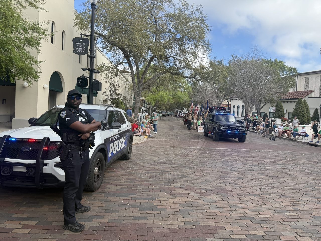 A police officer stands by a police SUV blocking a brick street, as people sit along the sidewalks and a parade with flags and vehicles approaches in the background.