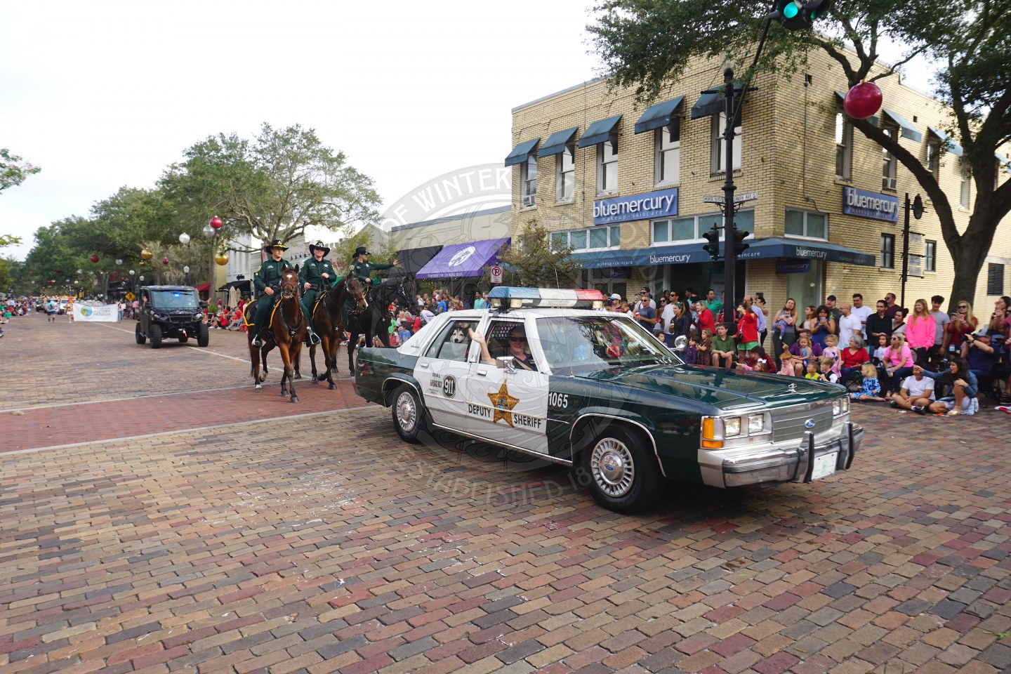 An antique police car with 2 people in front of the car and a black and white husky in the backseat with the window down. There are 3 sherriff deputies riding on horse behind the the antique police car.