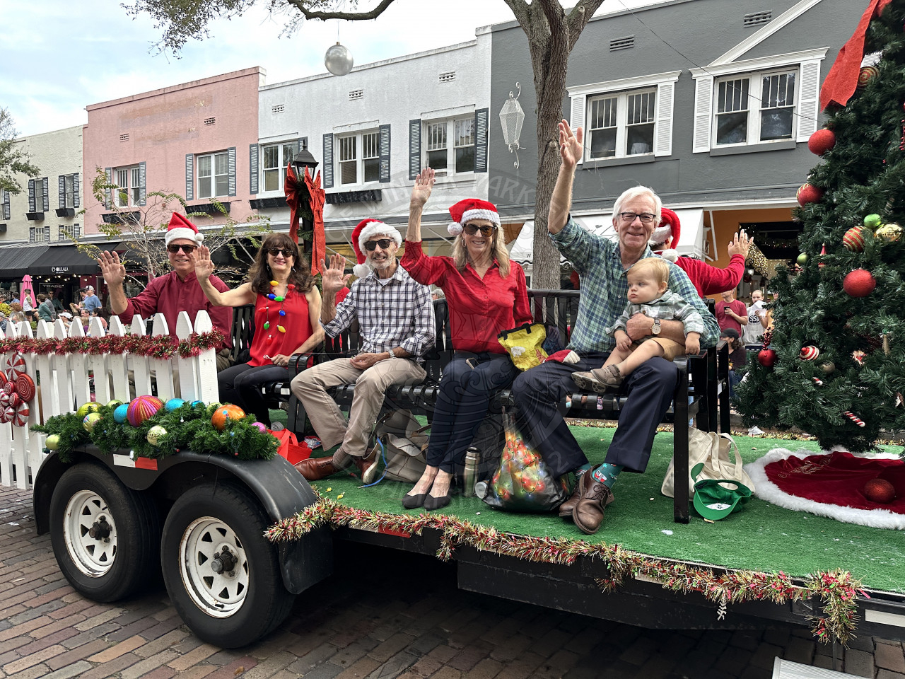 Commissioner Todd Weaver, Commissioner Sheila DeCiccio and her husband, and Mayor Anderson holding a baby in his lap with his wife sitting on a bench on the christmas parade float waving to the people sitting on the sidewalks watching them drive by.
