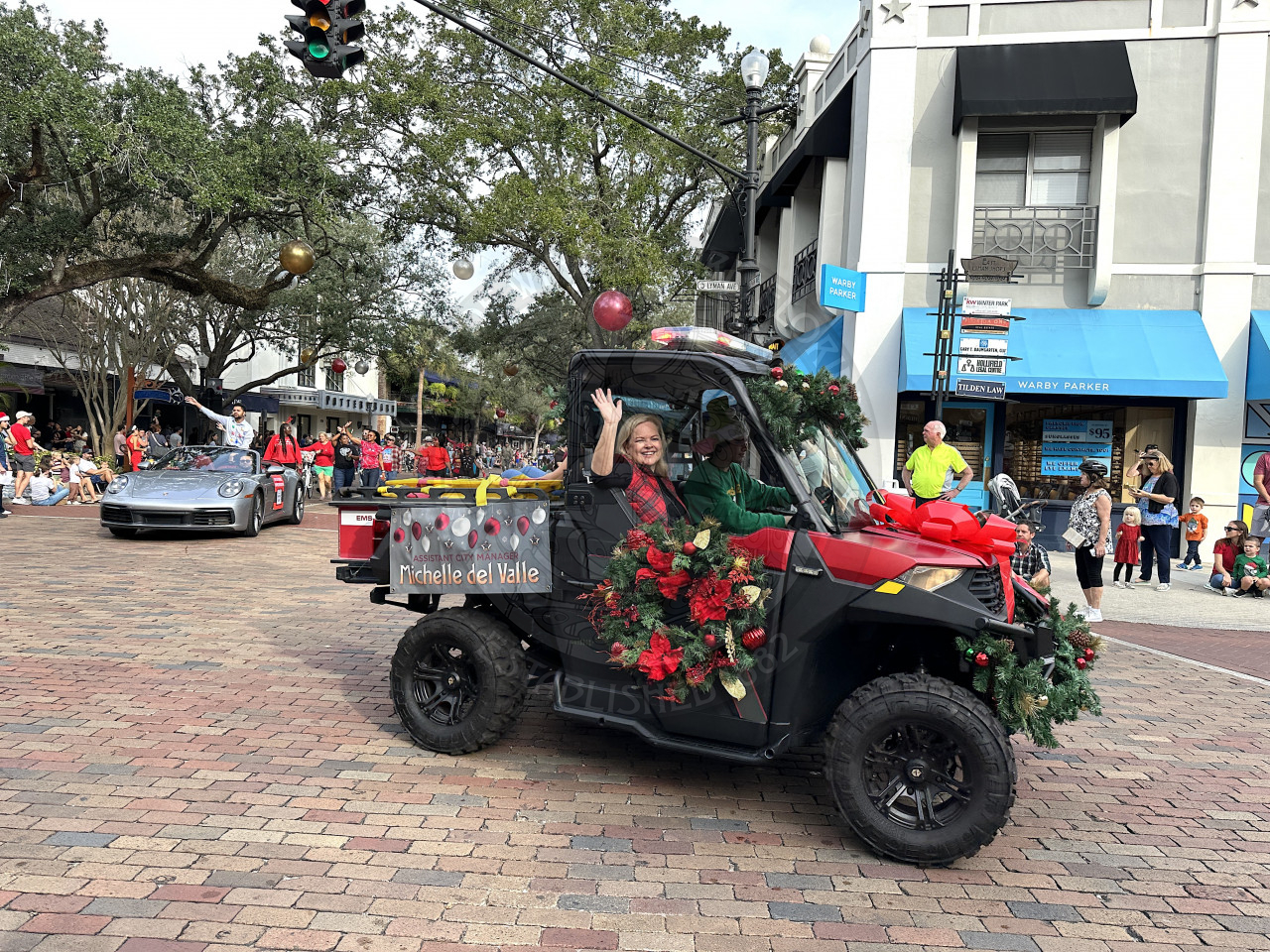 Assistant city manager, Michelle Del Valle sitting in the passenger seat waving to the crowd of peopleof an all-terrain vehicle that is decorated with a big red bow, garland and wreaths as they drive on Park Avenue. There is a banner on the back of the ATV that states her name and position. 
