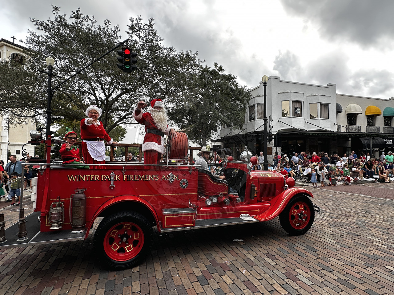 Santa claus waving and Mrs. Claus pointing to the crowd while they are standing on an antique 