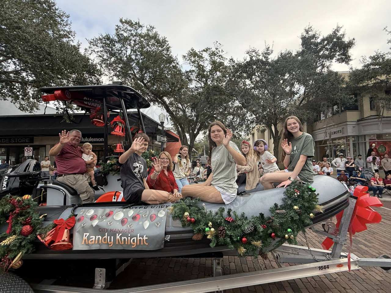 A family of grandparents and grandkids on a boat that is rolling on a brick road for a parade with people watching.