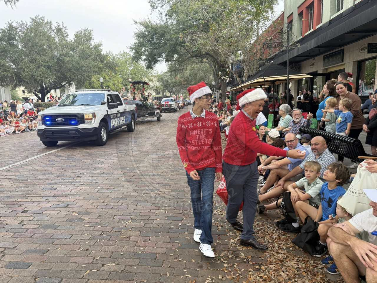 A son and a dad walking on a brick road as part of a parade passing out candy to people sitting on the sidewalk watching floats.