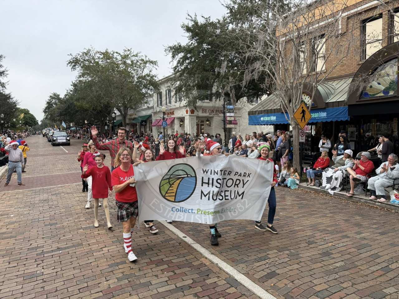 A group of people walking in a parade while holding a sign that says "Winter Park History Museum. Collect. Preserve. Exhibit."