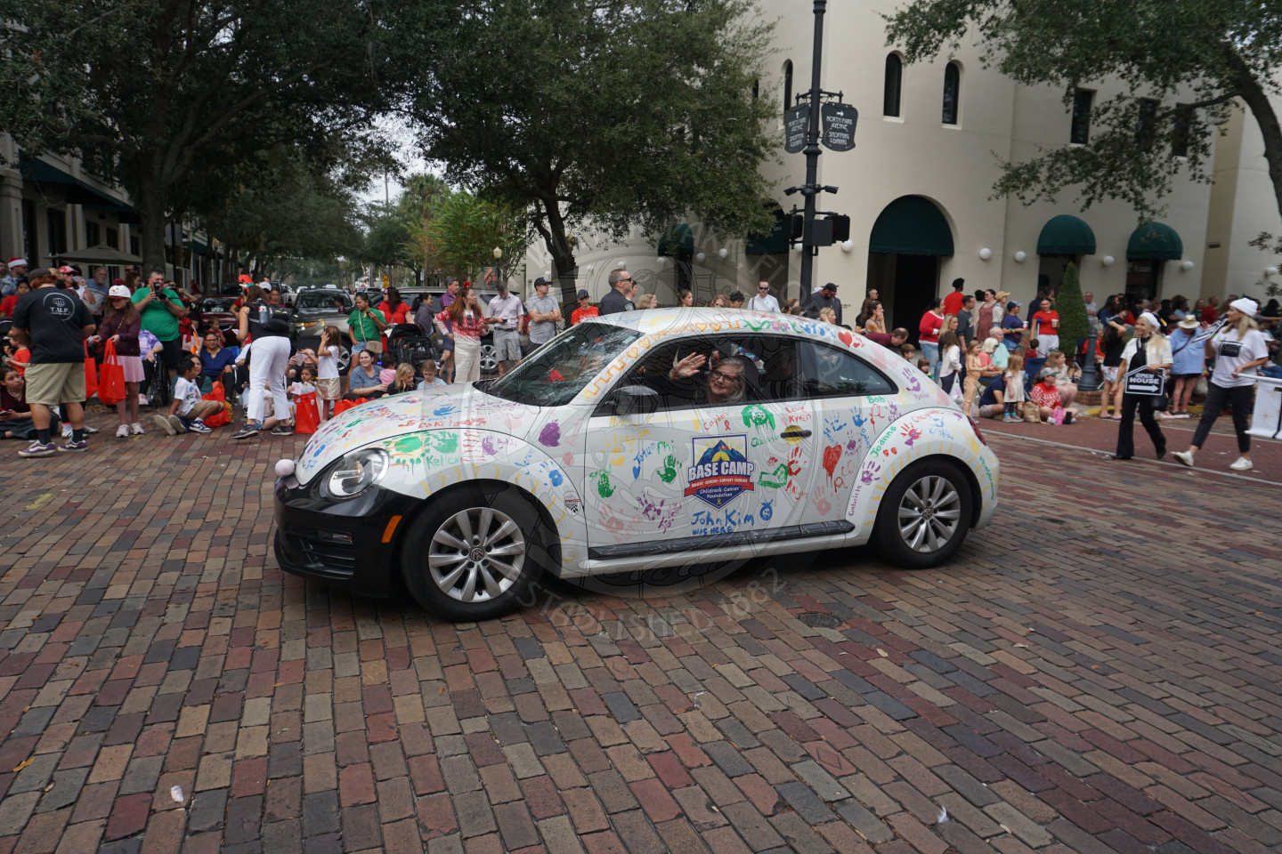 A white volkswagon with handprints all over it with the logo "Base Camp" ridding along a brick road in parade route.