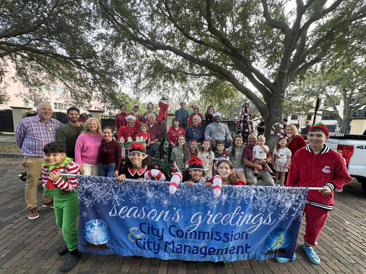 City management, commissioners and family all posing for a photo with a sign that says "Season's Greetings City Commission and City Management"