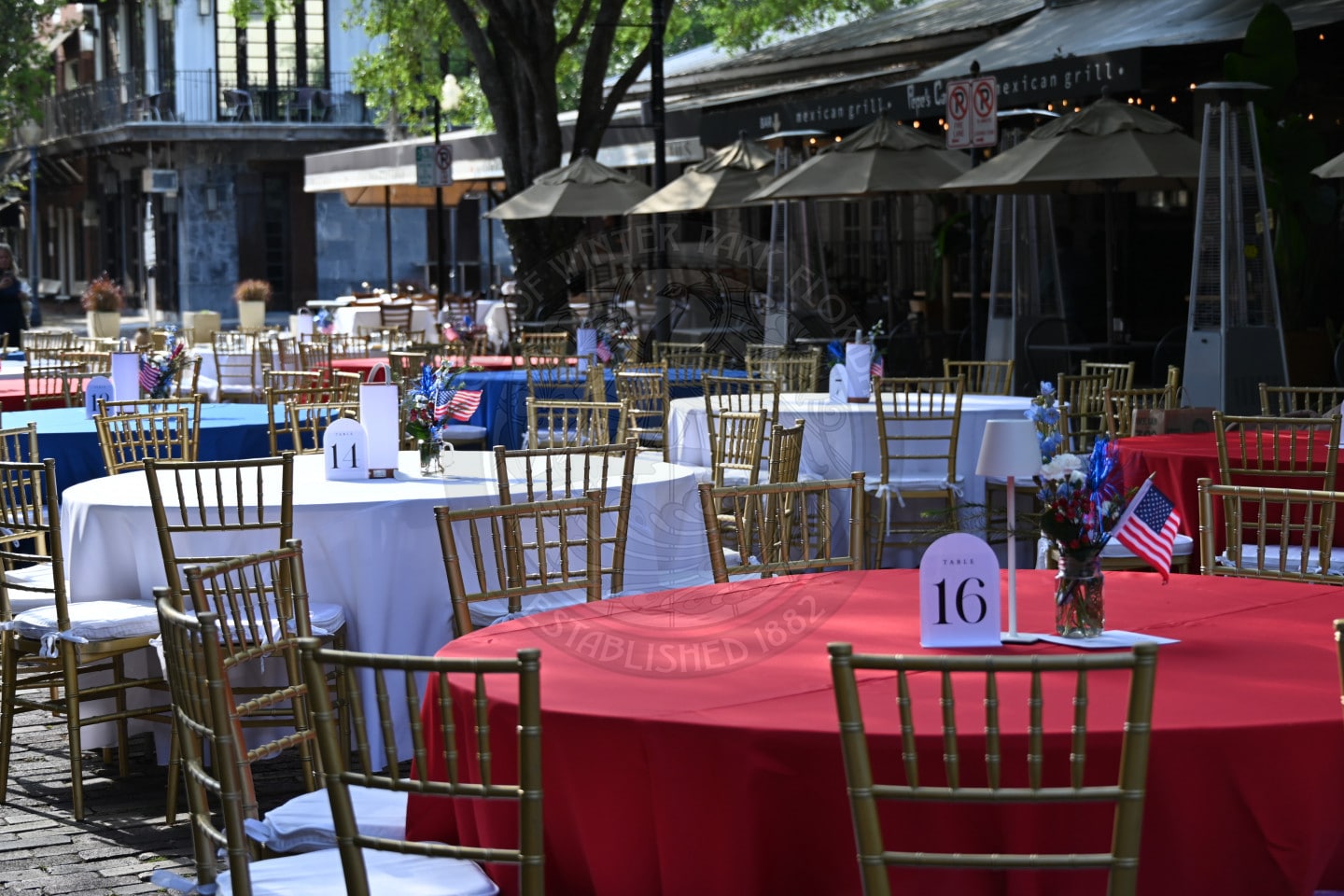 Outdoor dining area with tables covered in red, white, and blue cloths, decorated with small American flags.