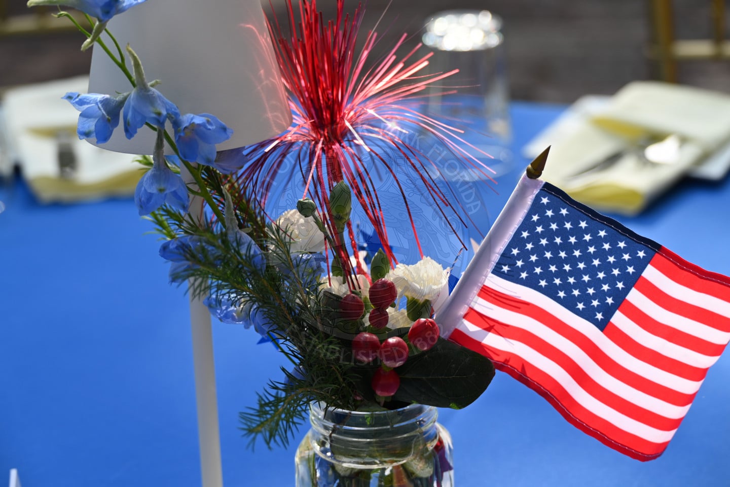 A mason jar centerpiece with an American flag, red and blue decorations, and flowers on a blue tablecloth.
