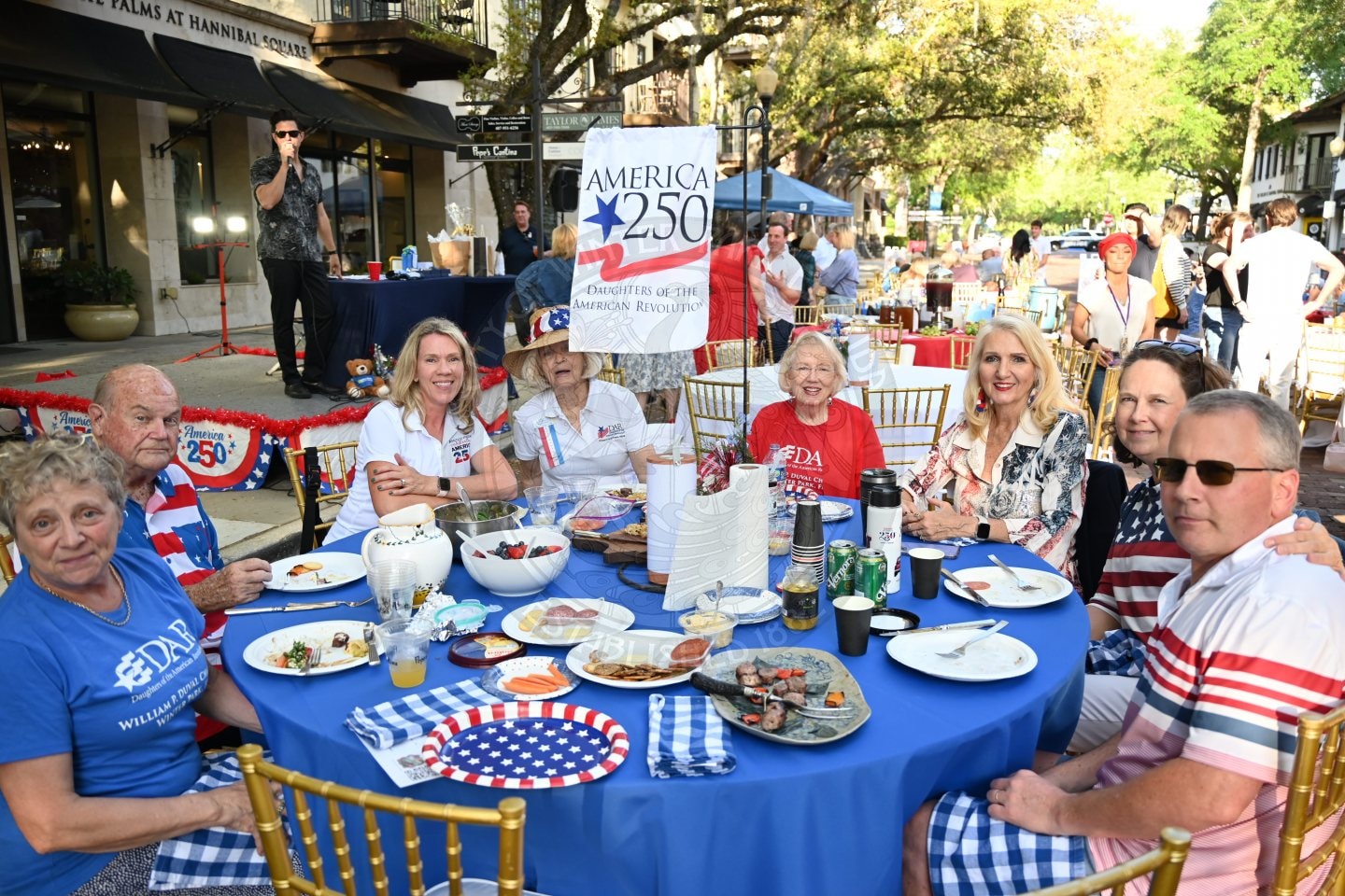A group of people in patriotic attire sit at a table with food, celebrating America 250 outdoors.
