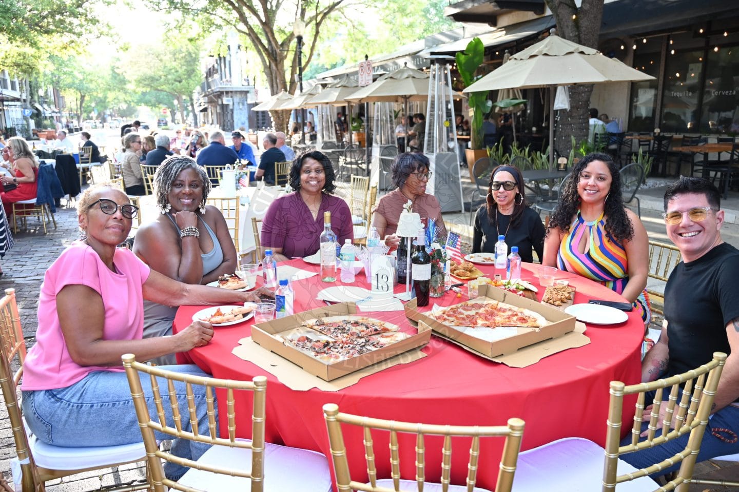 Seven people sit around a red table outdoors, smiling and sharing pizza on a sunny day with others dining nearby.