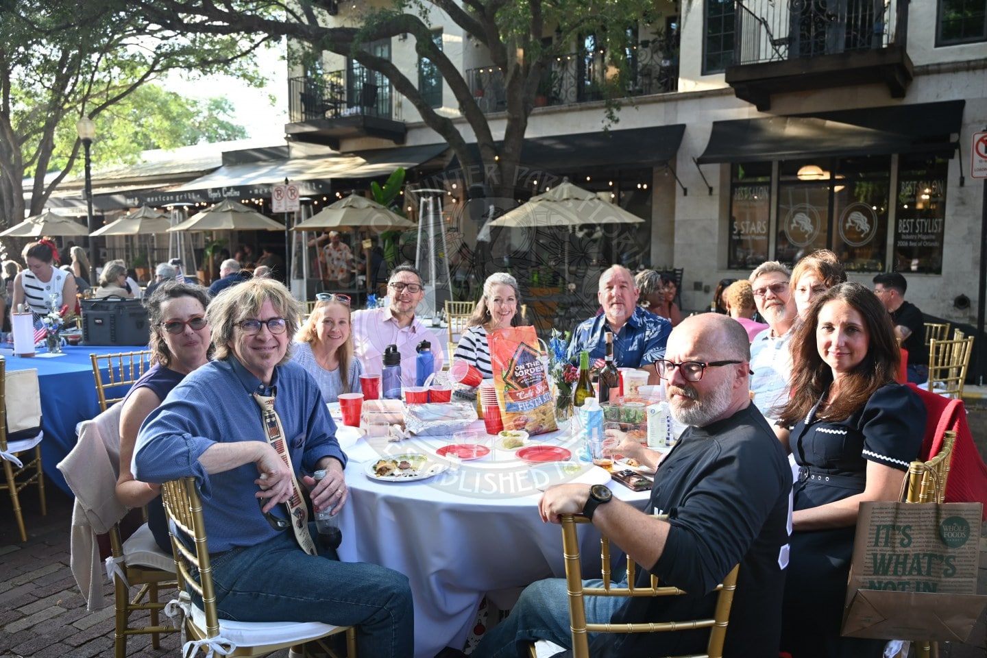A group of people sit around a table outdoors, smiling at a casual gathering with food and drinks.